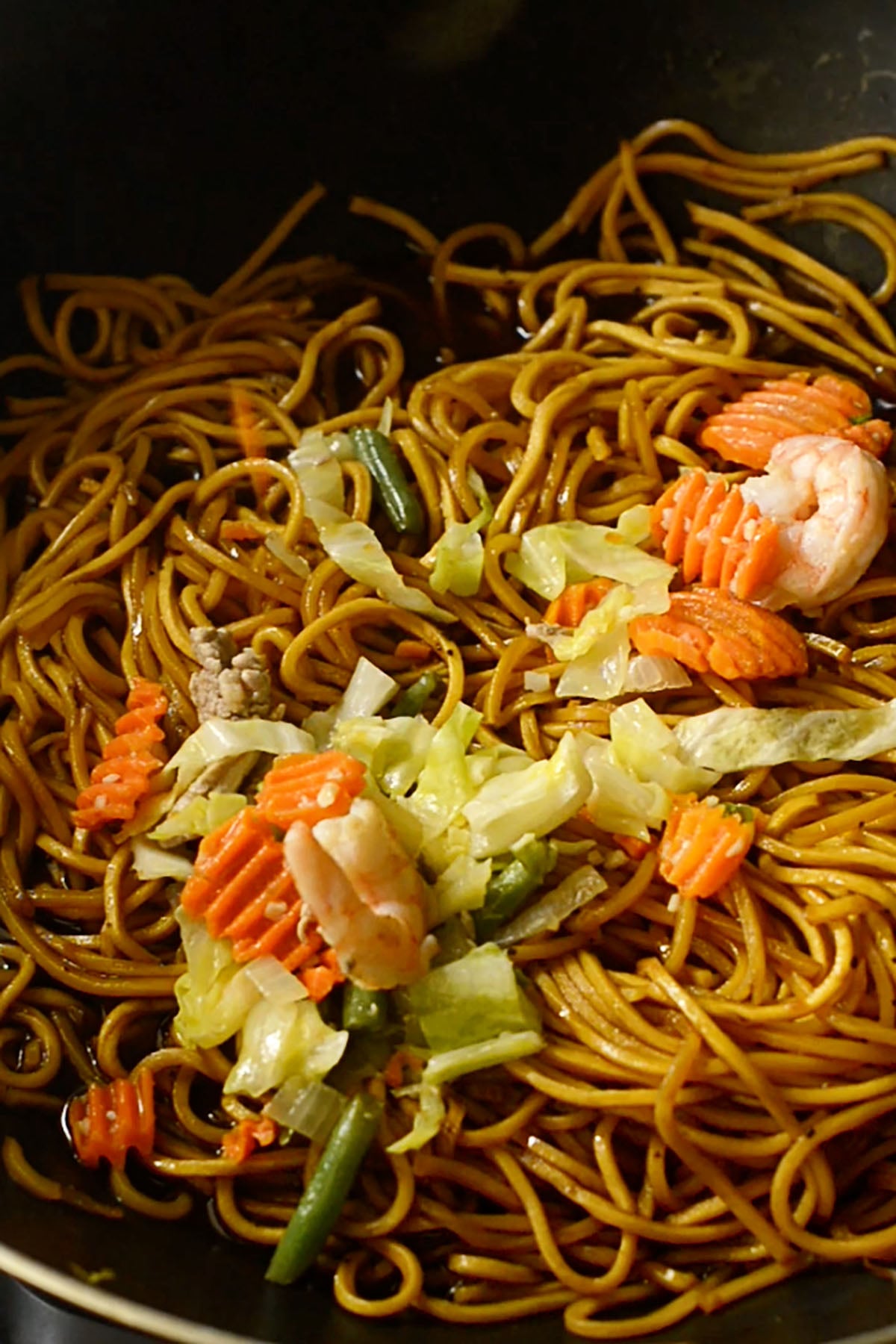 A cooked vegetable, shrimp and pork mixture being added to cooking pancit canton noodles.
